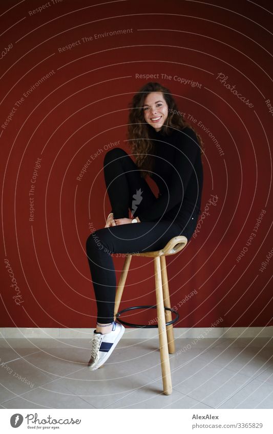 Young, tall woman sitting in front of a red wall on a wooden stool and smiling Lifestyle Style Joy already Living or residing Stool Young woman