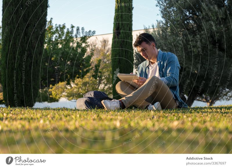 Student with backpack writing on notepad in park on bright day student grass education studying university casual learning young adult male academic working