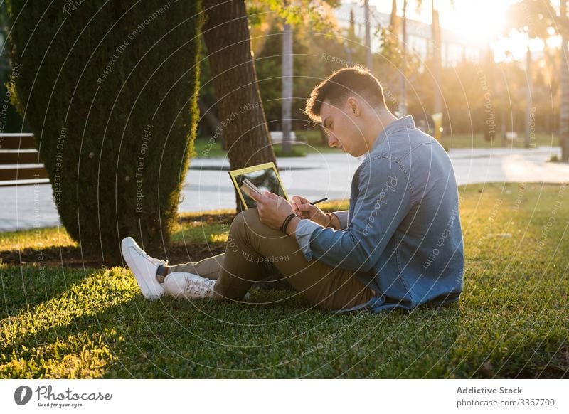 Student with backpack typing on laptop and notepad in park on bright day student grass computer education using studying university casual learning modern young