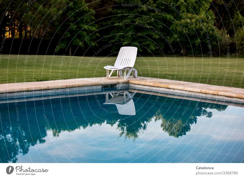 Corona thoughts | Stay at home - lonely deckchair stands at the edge of the pool, the sky is reflected in the water, a well-tended lawn and a row of trees can be seen in the background, the sun is setting.