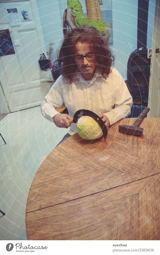 man sits at the kitchen table and tries to cut a cabbage with a sickle, with a hammer lying next to it, if this should prove too difficult ... Man cake Table