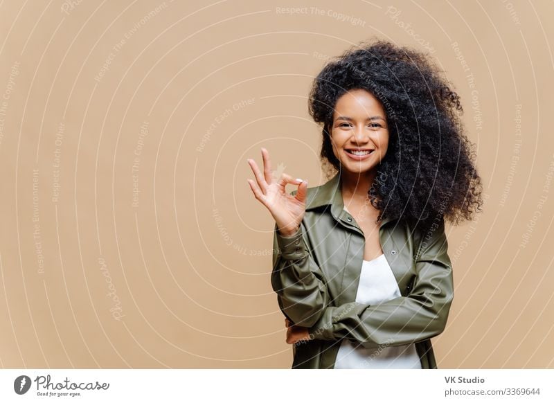 Indoor shot of pleasant looking curly woman has pleasant smile, makes okay gesture, excellent sign, gives approval, dressed in fashionable leather shirt, isolated over brown wall, blank space on left