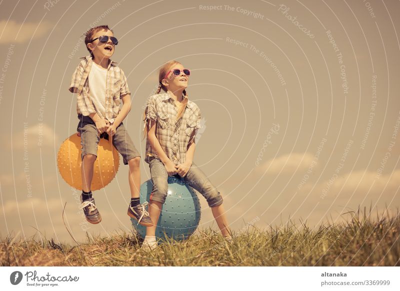 Brother and sister playing on the field at the day time. Children having fun outdoors. They jumping on inflatable balls on the lawn. Concept of friendly family.