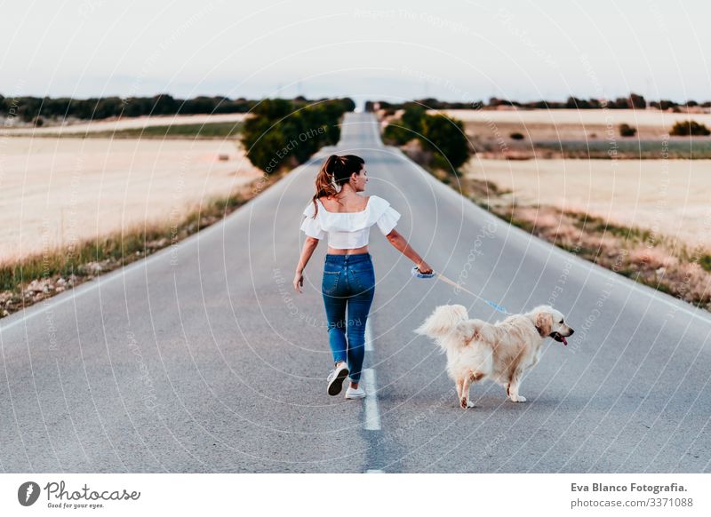 young woman walking by the road with her golden retriever dog at sunset. Pets outdoors street urban countryside field happy training labrador friendship park