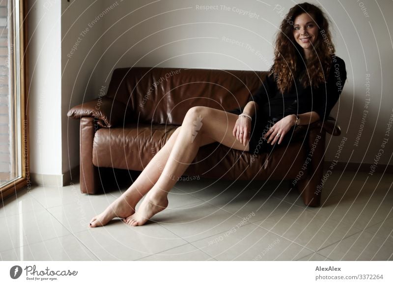 Portrait of a young, tall woman sitting barefoot on a brown leather couch and smiling Style Joy already Life Well-being Flat (apartment) Sofa Young woman