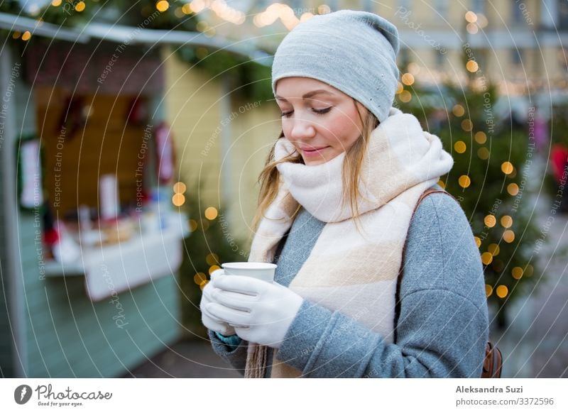 Young woman in Christmas market drinking cup of hot chocolate with marshmallow wearing knitted warm hat and scarf. Illuminated and decorated fair kiosks and shops on background. Helsinki, Finland