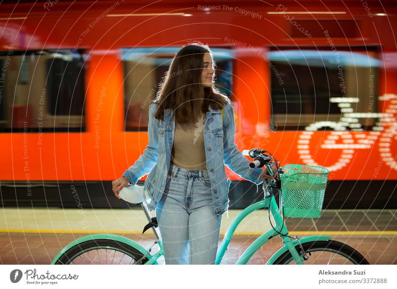 Teenager girl in jeans with yellow backpack and bike standing on metro station, waiting for train, smiling, laughing. Orange train passing by behind the girl. Futuristic subway station. Finland, Espoo