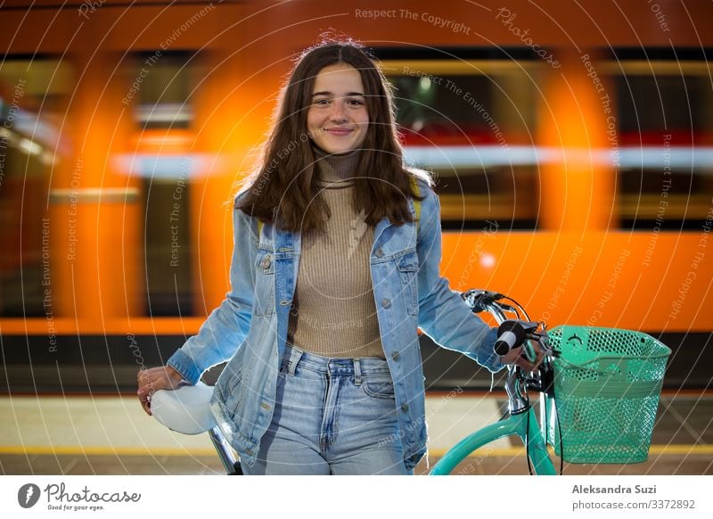 Teenager girl in jeans with yellow backpack and bike standing on metro station, waiting for train, smiling, laughing. Orange train passing by behind the girl. Futuristic subway station. Finland, Espoo