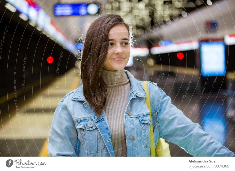 Teenager girl in jeans with backpack standing on metro station holding smart phone in hand, scrolling and texting, smiling and laughing. Futuristic bright subway station. Finland, Espoo