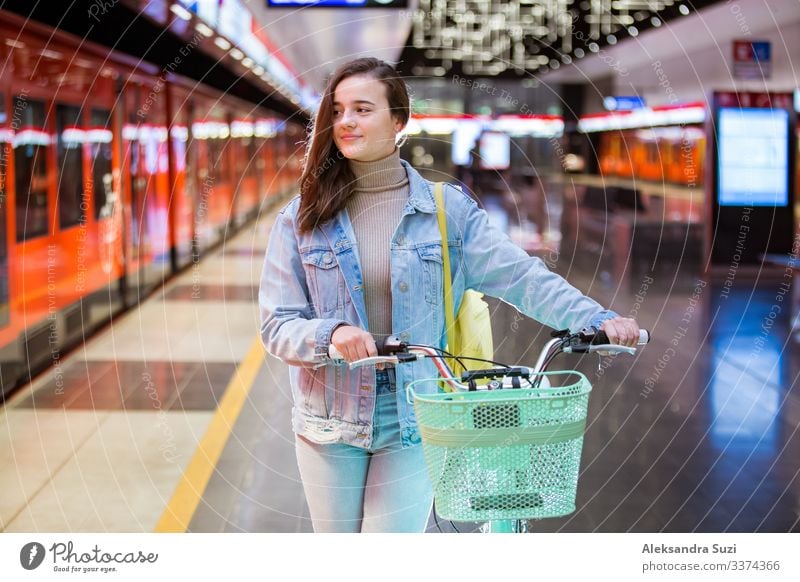 Teenager girl with backpack and bike standing on metro station holding smart phone in hand, scrolling and texting, smiling and laughing. Futuristic bright subway station. Finland, Espoo