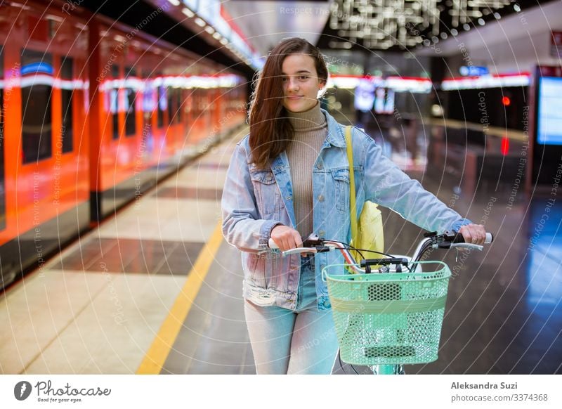 Teenager girl with backpack and bike standing on metro station holding smart phone in hand, scrolling and texting, smiling and laughing. Futuristic bright subway station. Finland, Espoo