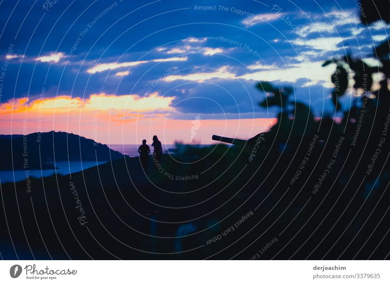 Two women talking at sunset on an island. Dark foreground with cannon. Blue clouds and with a view of a neighboring island. The women are only recognizable as shadows.