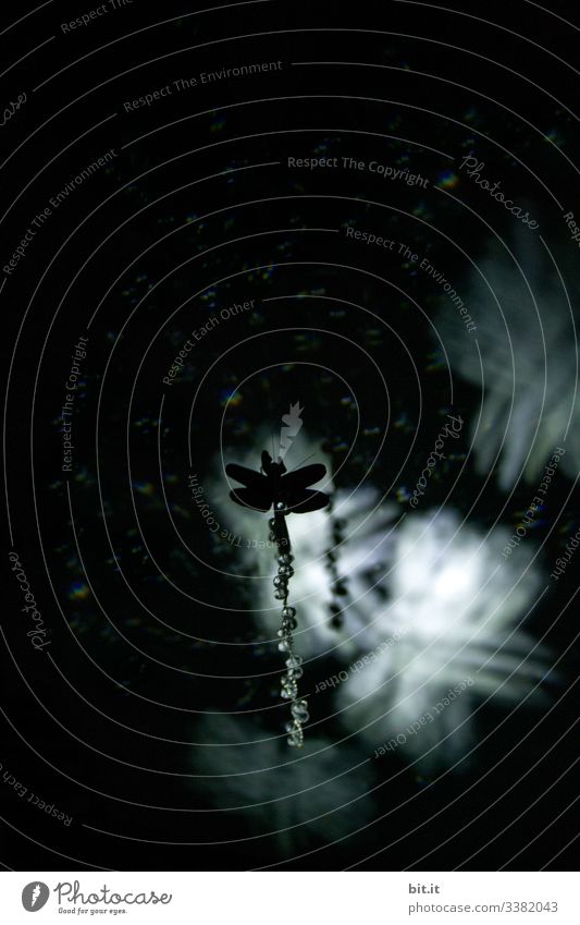 Silhouette of a dragonfly made of clay paper floats on a chain, in front of a black background with luminous dragonflies, in black and white, with light and shadow.