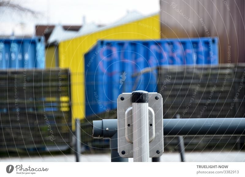 Behind an elegant white and grey barrier old wooden pallets and two blue containers are waiting in front of a yellow warehouse and brown facade Container