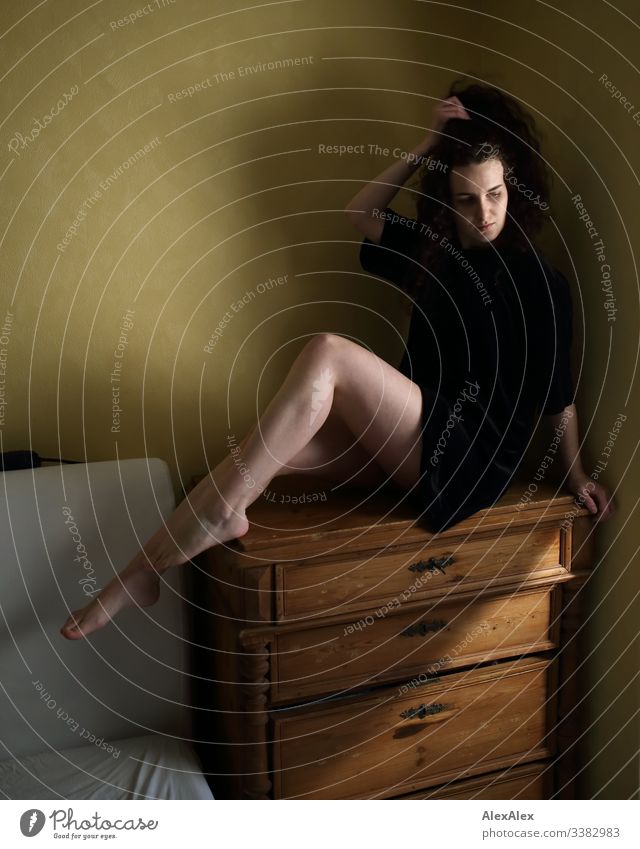 Young woman sitting on a chest of drawers in front of a yellow wall Athletic beautiful woman Looking into the camera portrait Central perspective