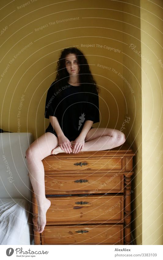 Young woman sitting on a chest of drawers in front of a yellow wall Looking into the camera portrait Central perspective Shallow depth of field Day