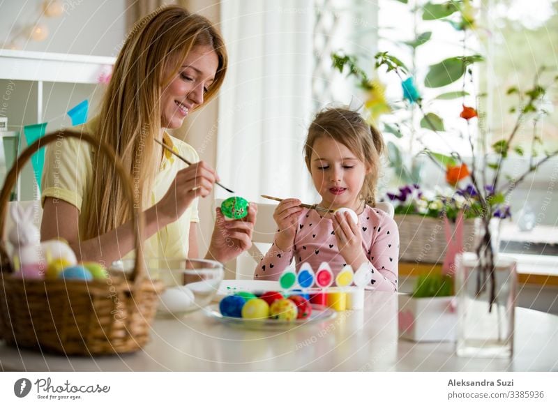 A mother and daughter celebrating Easter, painting eggs with brush. Happy family smiling and laughing. Cute little girl in bunny ears preparing the holiday.