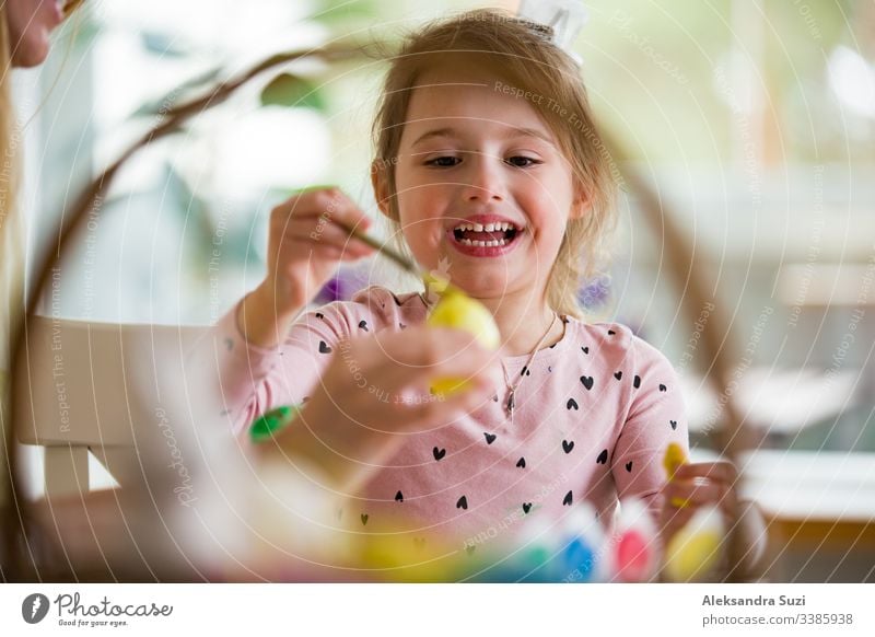 A mother and daughter celebrating Easter, painting eggs with brush. Happy family smiling and laughing. Cute little girl in bunny ears preparing the holiday.