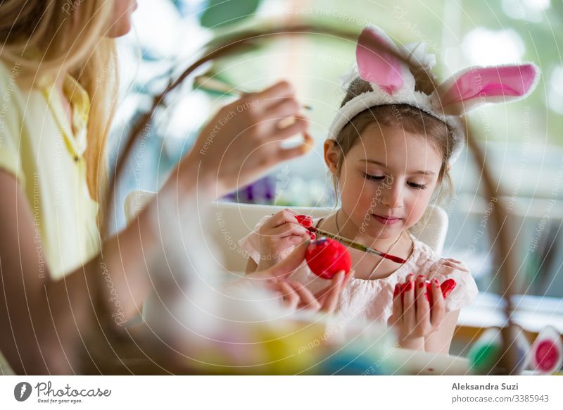 A mother and daughter celebrating Easter, painting eggs with brush. Happy family smiling and laughing. Cute little girl in bunny ears preparing the holiday.