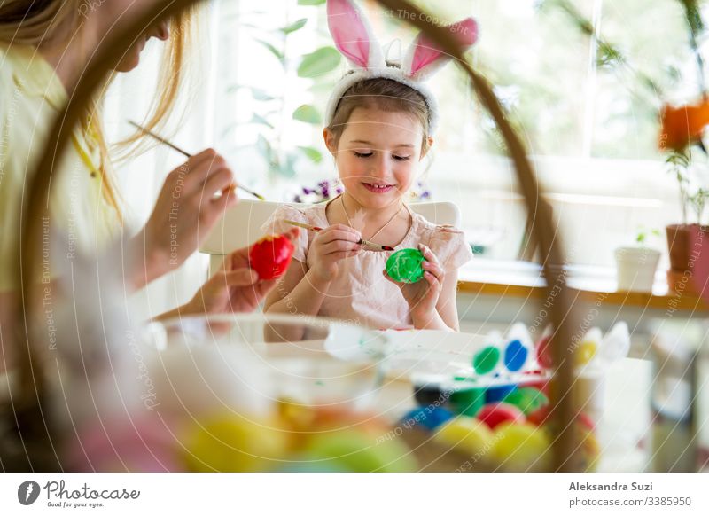 A mother and daughter celebrating Easter, painting eggs with brush. Happy family smiling and laughing. Cute little girl in bunny ears preparing the holiday.