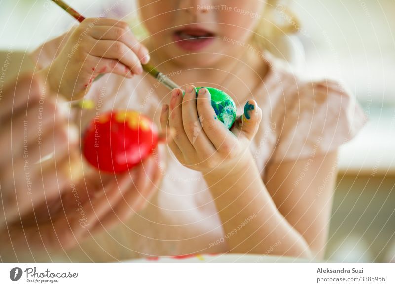 A mother and daughter celebrating Easter, painting eggs with brush. Happy family smiling and laughing. Cute little girl in bunny ears preparing the holiday.