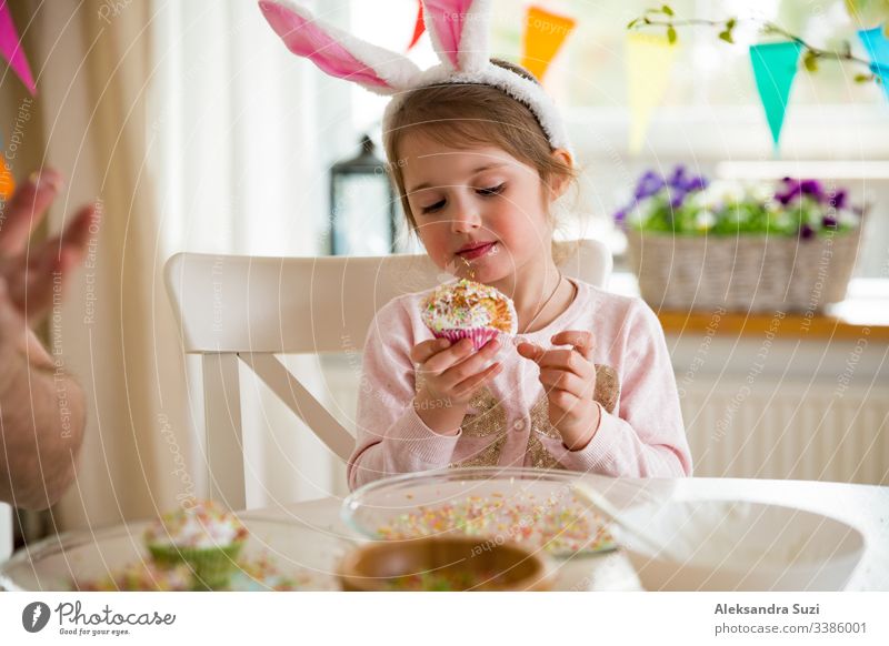 Father and daughter celebrating Easter, eating cupcakes covered with glaze. Happy family holiday. Cute little girl in bunny ears. Beautifully decorated room