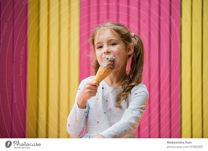 Cute little girl eating chocolate ice cream. Smiling and laughing. Colorful pink and yellow wall on background. Bright summer concept beautiful bright cafe