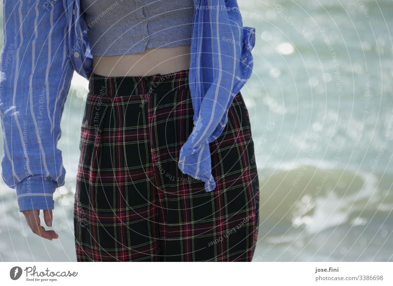 Young woman, neckline upper body and legs in front of the sea Shirt Checkered Striped Showing one's bellybutton windy Hand Waves Blue Cloth Detail Nature