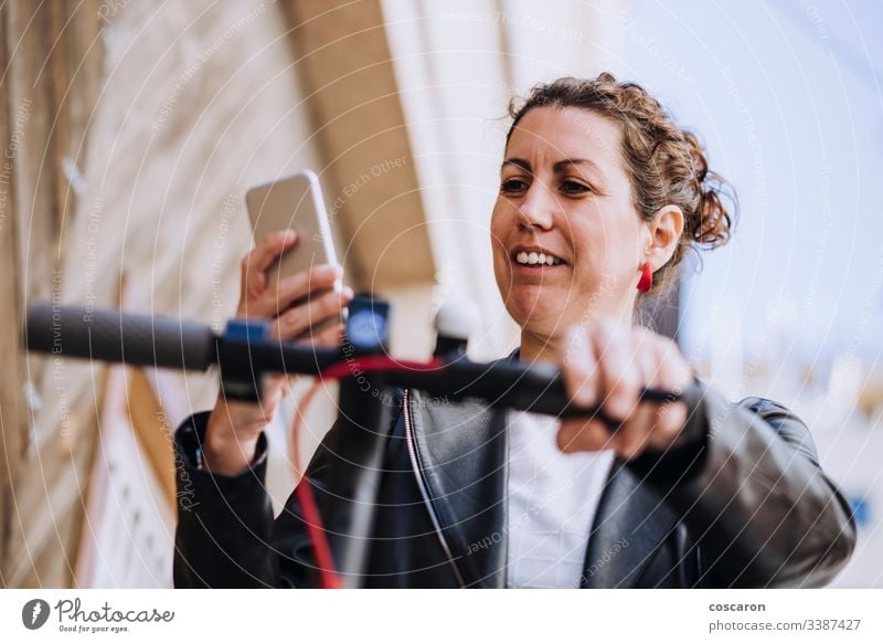 Middle aged woman riding his electric scooter and looking at mobile phone. active activity app autumn background beautiful bike blond booking cars city