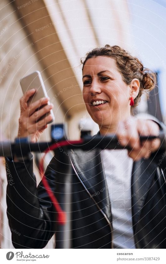 Middle aged woman riding his electric scooter and looking at mobile phone. active activity app autumn background beautiful bike blond booking cars city