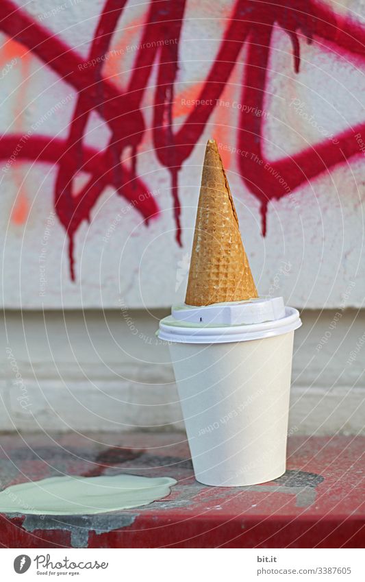 Ice leaves a mess. Empty ice cream wafer lies lastingly, upside down on an empty white coffee mug from a coffee to go. Colourful graffiti decorates the wall in the background.