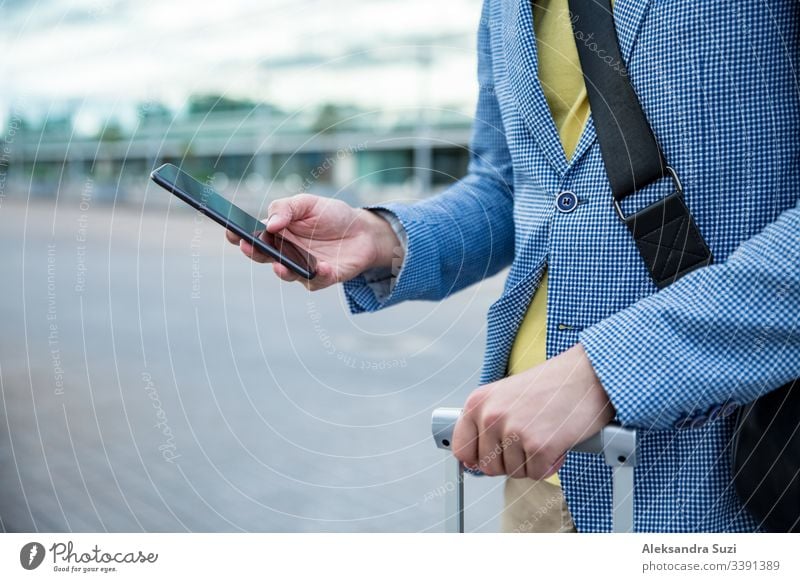 Stylish man standing at airport with smartphone and suitcase, browsing, texting, using mobile app. Business traveling. application arrival bag business