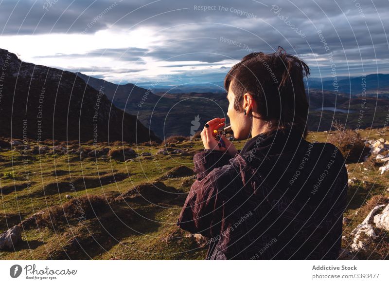 Cool young woman lighting cigarette standing against amazing landscape of mountain ridge in cloudy weather highland smoke rebel travel cool escape hipster fire