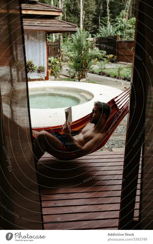 Young male tourist in swimwear with book on hammock man resort hotel lounge chill poolside read exotic tropical swimsuit relax rest sit lifestyle young enjoy