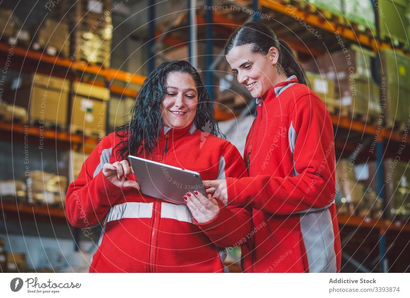 Satisfied smart female colleagues browsing digital tablet warehouse women using search watch device smile enjoy gadget storage logistic modern together work