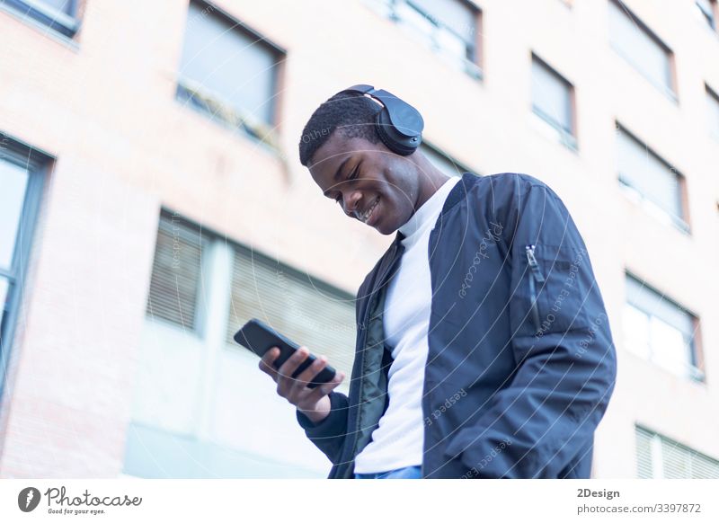 Young Black man standing in the street while listening music by headphones technology lifestyle male black casual attire trendy happy african earphones guy