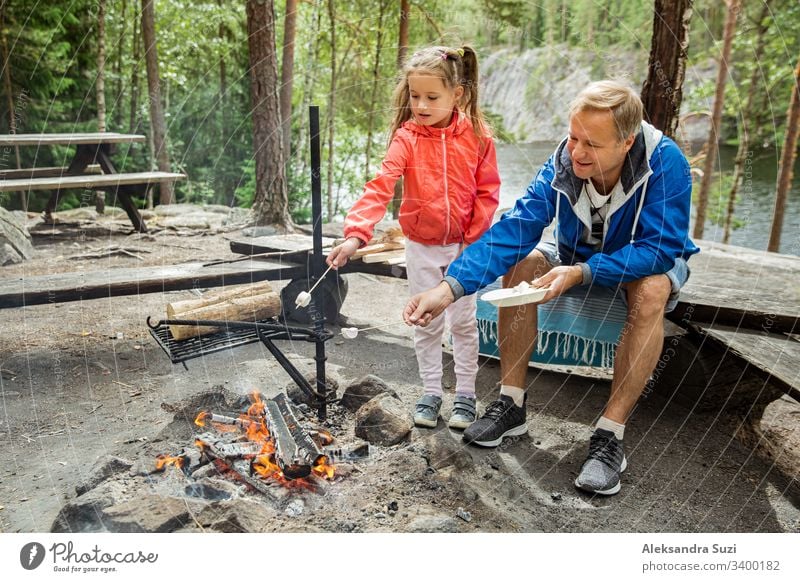 Man and his little daughter having barbecue in forest on rocky shore of lake, making a fire, grilling bread, vegetables and marshmallow. Family exploring Finland. Scandinavian summer landscape.