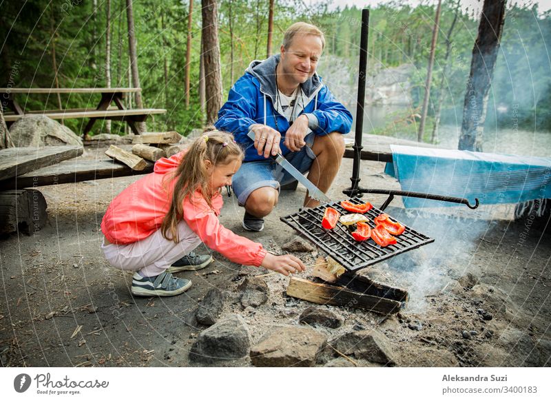 Man and his little daughter having barbecue in forest on rocky shore of lake, making a fire, grilling bread, vegetables and marshmallow. Family exploring Finland. Scandinavian summer landscape.