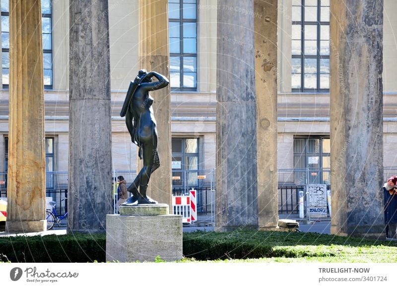 Naked woman of bronze stands on a pedestal between columns in the sun and ties a quiver with arrows on her back: Diana, the goddess of the hunt. In the background the sky is reflected in large windows of a building.