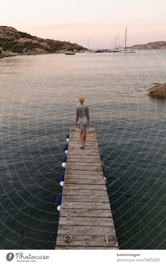 Beautiful woman in luxury summer dress standing on wooden pier enjoying peaceful seascape at dusk. Female traveler stands on a wooden pier in Porto Rafael, Costa Smeralda, Sardinia, Italy.