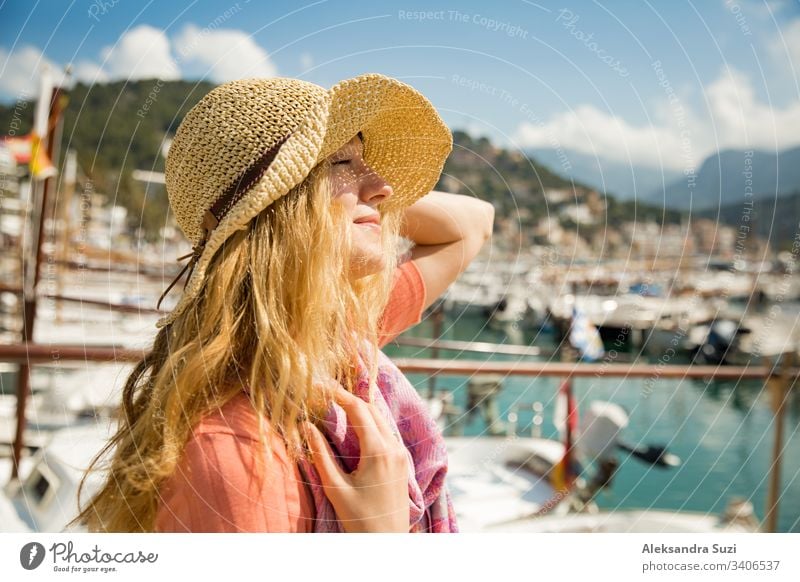 Portrait of young woman with light curly hair in straw hat enjoying sun and breeze, smiling. Sunny harbor with boats and yachts, green mountains on background. Enjoying life, happy person traveling,