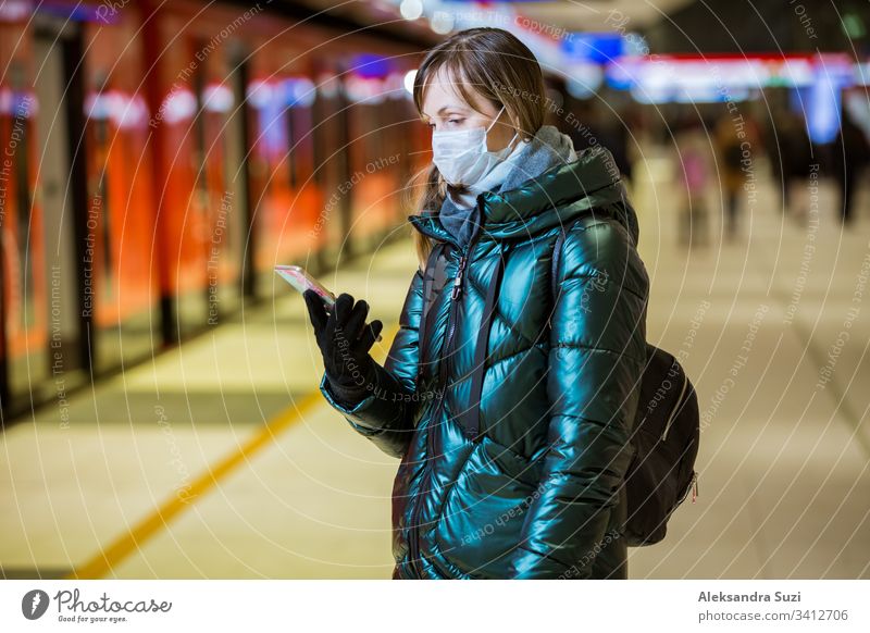 Woman in winter coat with protective mask on face standing on metro station, waiting for train, looking worried. Preventive measures in public places of epidemic regions. Finland, Espoo