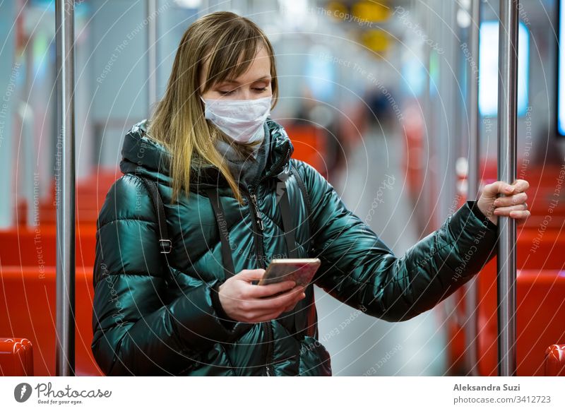 Woman in winter coat with protective mask on face standing in subway car, using phone, looking worried. Preventive measures in public places of epidemic regions. Finland, Espoo