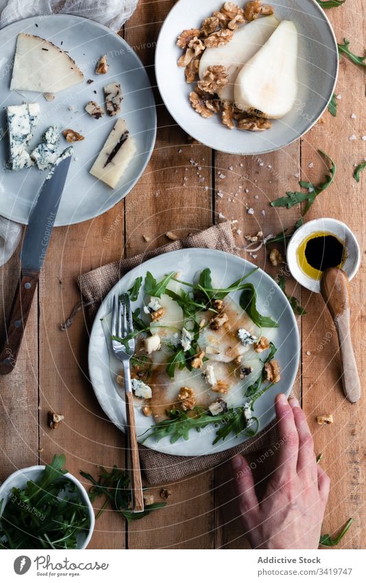 Crop person putting plate with salad near ingredients table rustic kitchen pear arugula walnut cheese delicious food tasty fresh dish gourmet meal cuisine