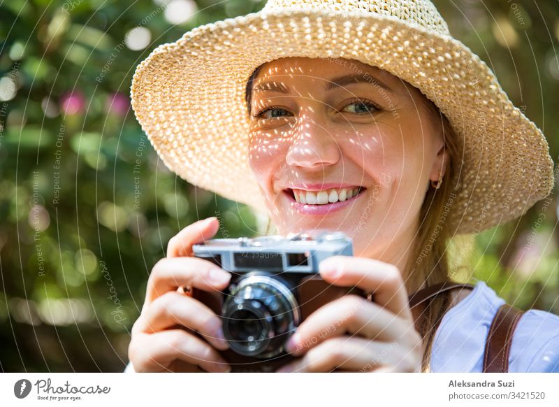 Close-up portrait of a beautiful woman in straw hat travelig in tropic forest, taking photos on retro camera. Light shadows through the cut-out detailed brim on face. Tourist with backpack.