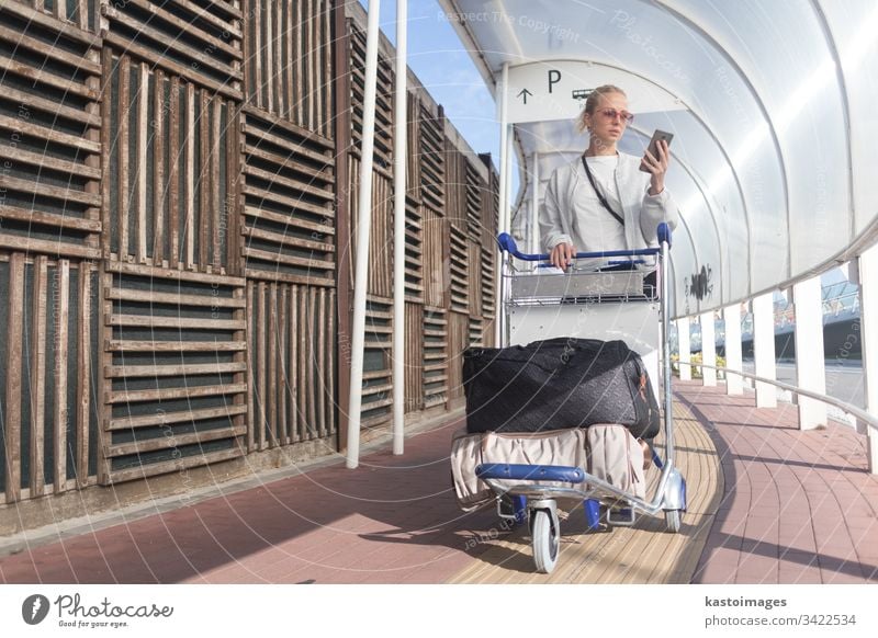 Young casual woman using mobile phone application while transporting luggage from arrival parking to international airport departure termainal by luggage trolley