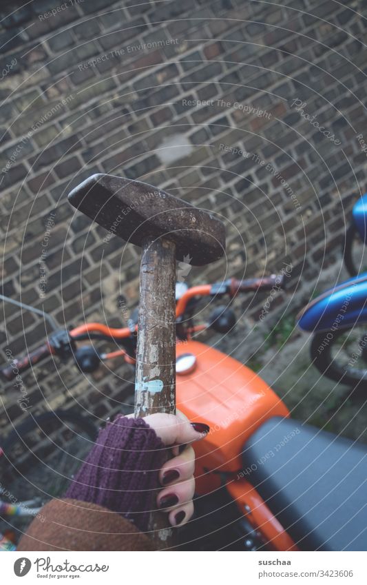 woman holding a hammer over an orange moped in front of a brick wall Woman feminine Hand Fingers Hammer Tool painted fingernails Motorcycle Handlebars Wheel