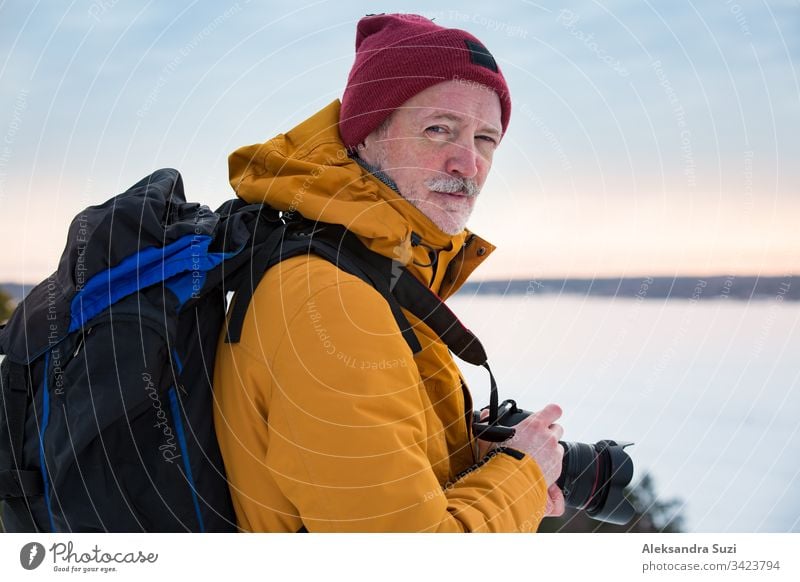 Portrait of mature man with grey beard exploring Finland in winter. Traveler Taking pictures on the top of rock. Beautiful view of northern landscape with frozen Baltic Sea and snowy islands.