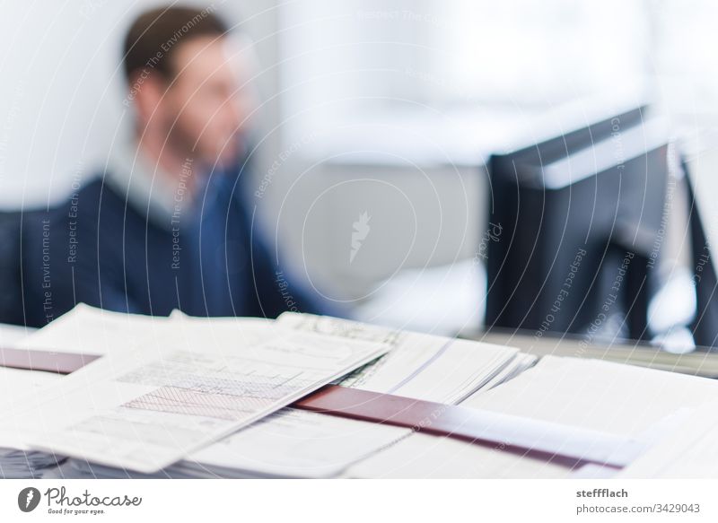 Architects working material in the foreground, in the background a blurred employee in front of the monitor laptop people Office office Phone Table Workplace