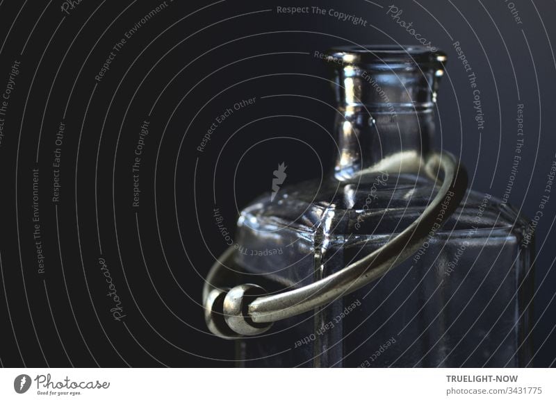 An old silver-plated bangle from India lies on an empty square glass bottle and these two objects reflect a little daylight in front of an almost black background, low key